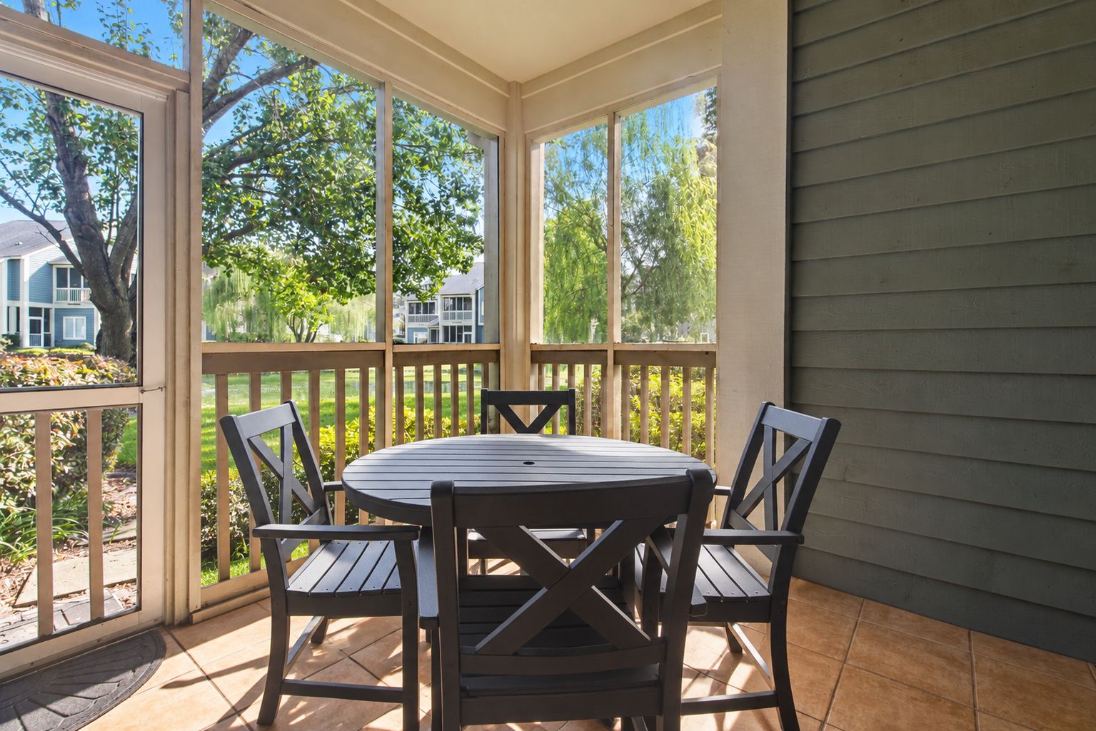A covered patio with a round table and four chairs, surrounded by a wooden fence and large windows.