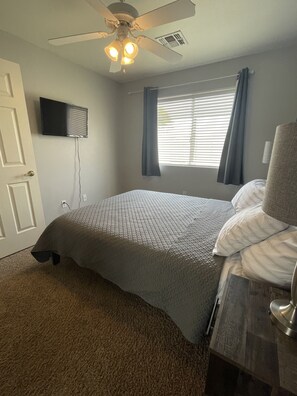 Une chambre à coucher comprenant un lit, un ventilateur de plafond, une fenêtre avec des rideaux, un téléviseur à écran plat fixé au mur et une porte.