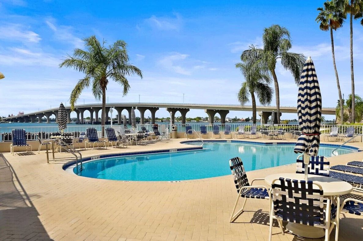 A swimming pool with lounge chairs and umbrellas, palm trees, and a bridge in the background.