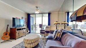 A living room with a sofa, ottoman, TV, guitar, and a balcony with chairs. - Bahia Vista 9-521 by Distinctive Beach Rentals (St. Petersburg)