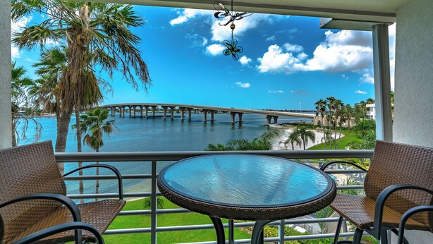 A balcony with wicker chairs, a glass table, and a view of a bridge over water with palm trees.