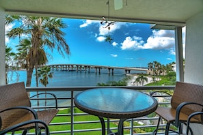 A balcony with wicker chairs, a glass table, and a view of a bridge over water with palm trees. - Bahia Vista 9-521 by Distinctive Beach Rentals (St. Petersburg)