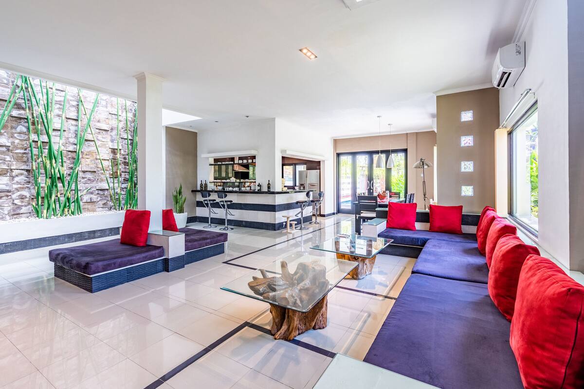 Amplia sala de estar con una mesa de centro de vidrio, sofás rojo y azul, y una cocina al fondo.