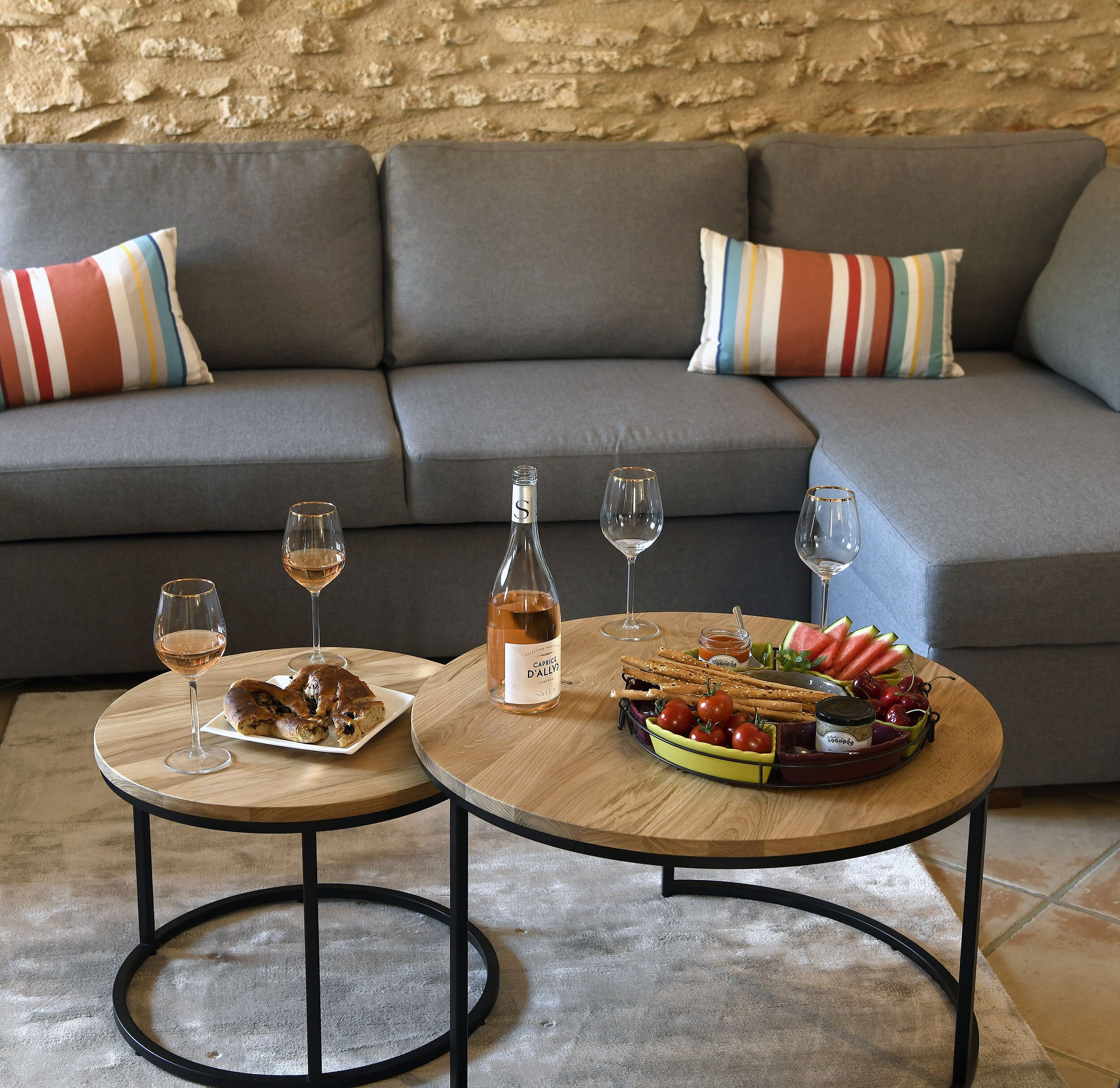 Un espacio de sala acogedor con un sofá gris, almohadones a rayas, dos mesas redondas de madera y una variedad de bocadillos y bebidas.