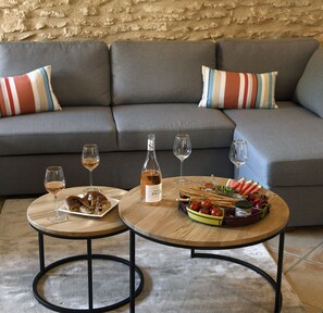 Ein gemütlicher Wohnbereich mit grauem Sofa, gestreifter Dekokissen, zwei runden Holztischen und einer Auswahl an Snacks und Getränken.