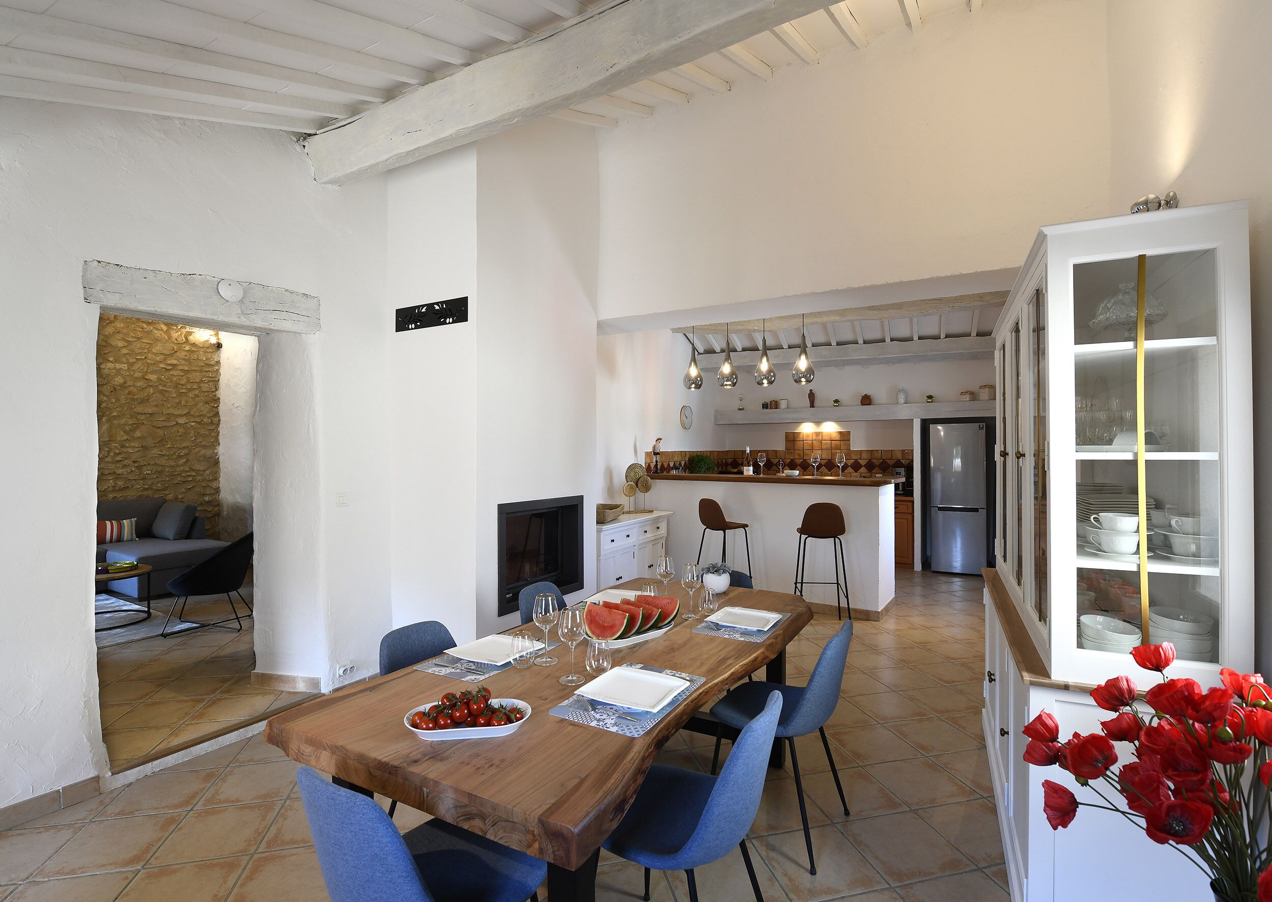 Comedor con una mesa de madera dispuesta para cuatro, sillas azules, una cocina al fondo y un aparador de vidrio con vajilla.