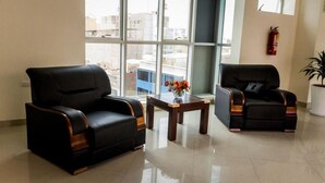 A modern hotel lobby with two black leather sofas, a small wooden table with a vase of flowers, and a large window offering a city view.