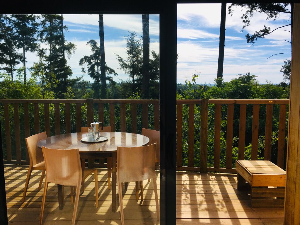 An outdoor wooden deck with a round table and chairs, surrounded by a wooden fence and dense greenery.