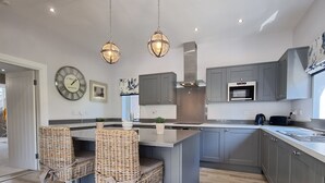 Una cocina moderna con gabinetes grises, una isla central, sillas de mimbre, iluminación colgante y un reloj grande en la pared.