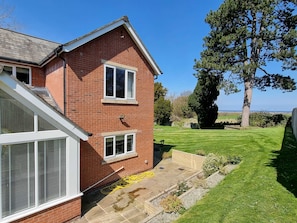 Une maison à deux étages, construite en briques, avec un toit en pente, un petit jardin devant avec une pelouse et un grand arbre.