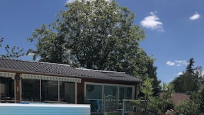 Una piscina con solárium empedrado, un porche cubierto y un árbol grande al fondo.