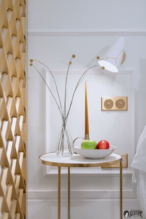 Un mur blanc orné d’un panneau décoratif, un vase blanc contenant des branches et un saladier rempli de fruits, ainsi qu’une lampe blanche.