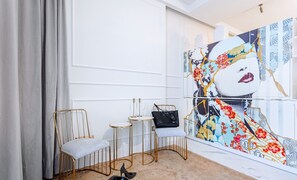 A modern interior with a seating area featuring two gold-framed chairs and a small round table, a black handbag on a chair, and a large abstract painting on the wall.