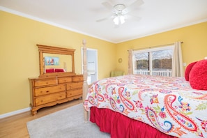 A bedroom with a wooden dresser, a bed with a colorful bedspread, a ceiling fan, and a window with a view.