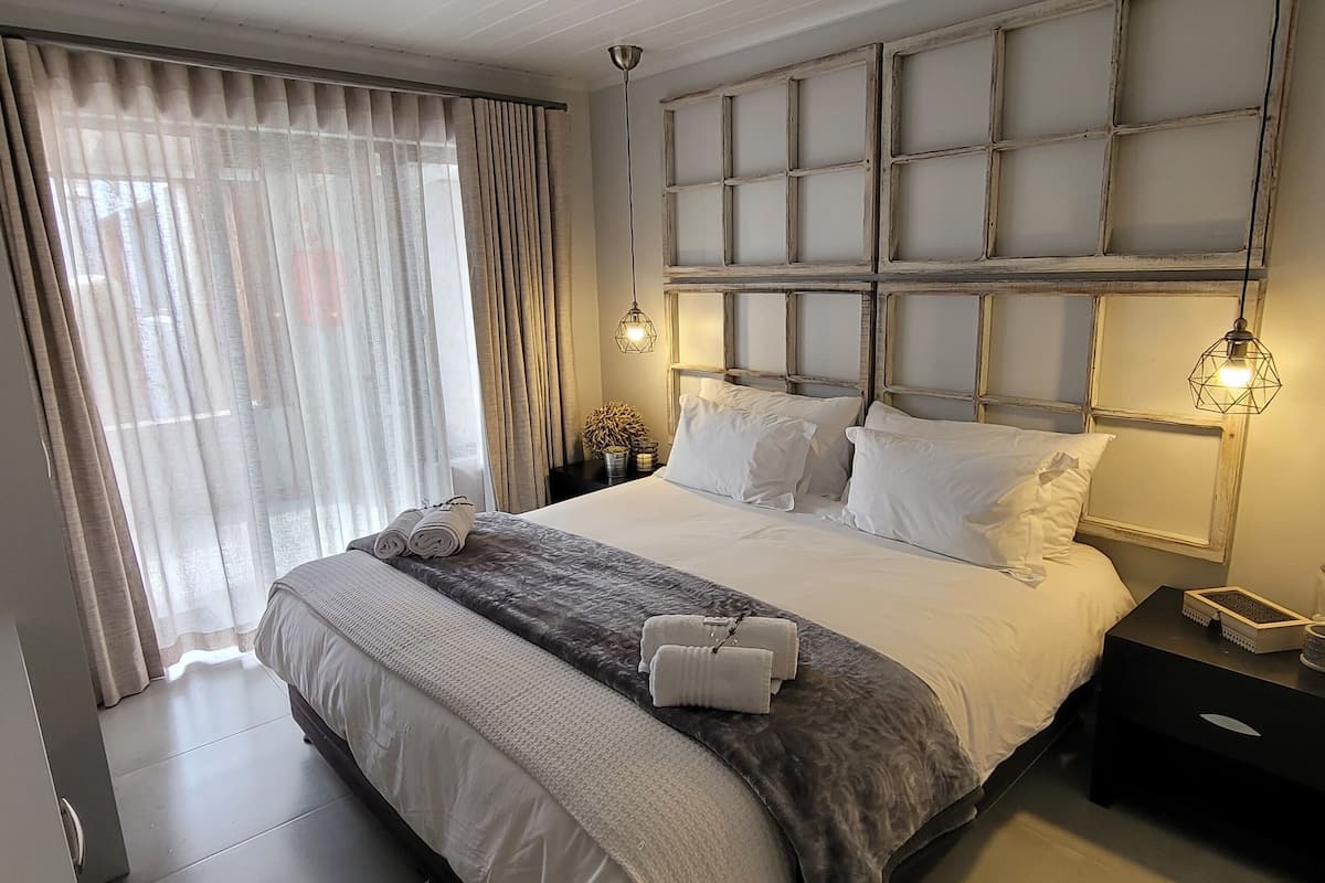 A neatly made bed with white linens and a gray throw blanket, a bedside table with a lamp, and a window with sheer curtains.