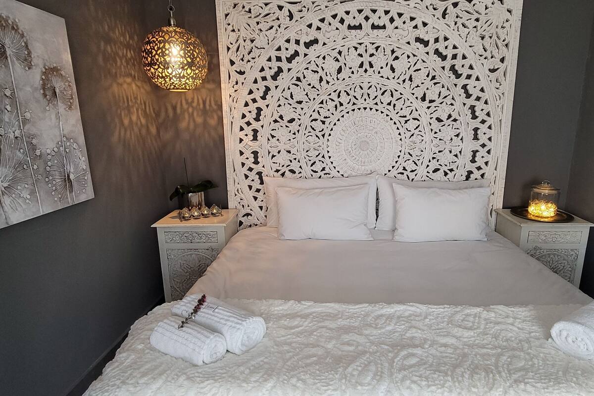A neatly made bed with white linens and towels, a decorative wall hanging, and a patterned headboard.