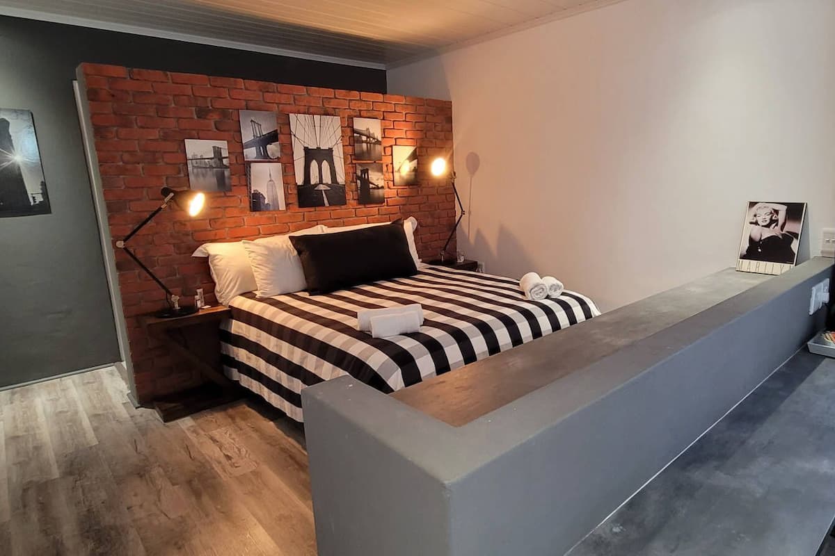 A modern hotel room with a brick accent wall, a bed with striped bedding, a desk with a chair, and a window with curtains.