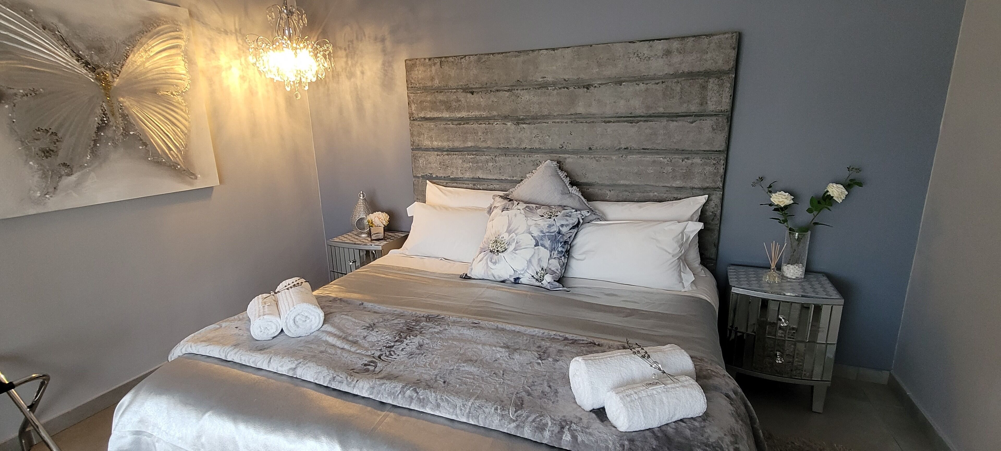 A bedroom with a large bed, a chandelier, a decorative wall piece, and a nightstand with a vase of flowers.