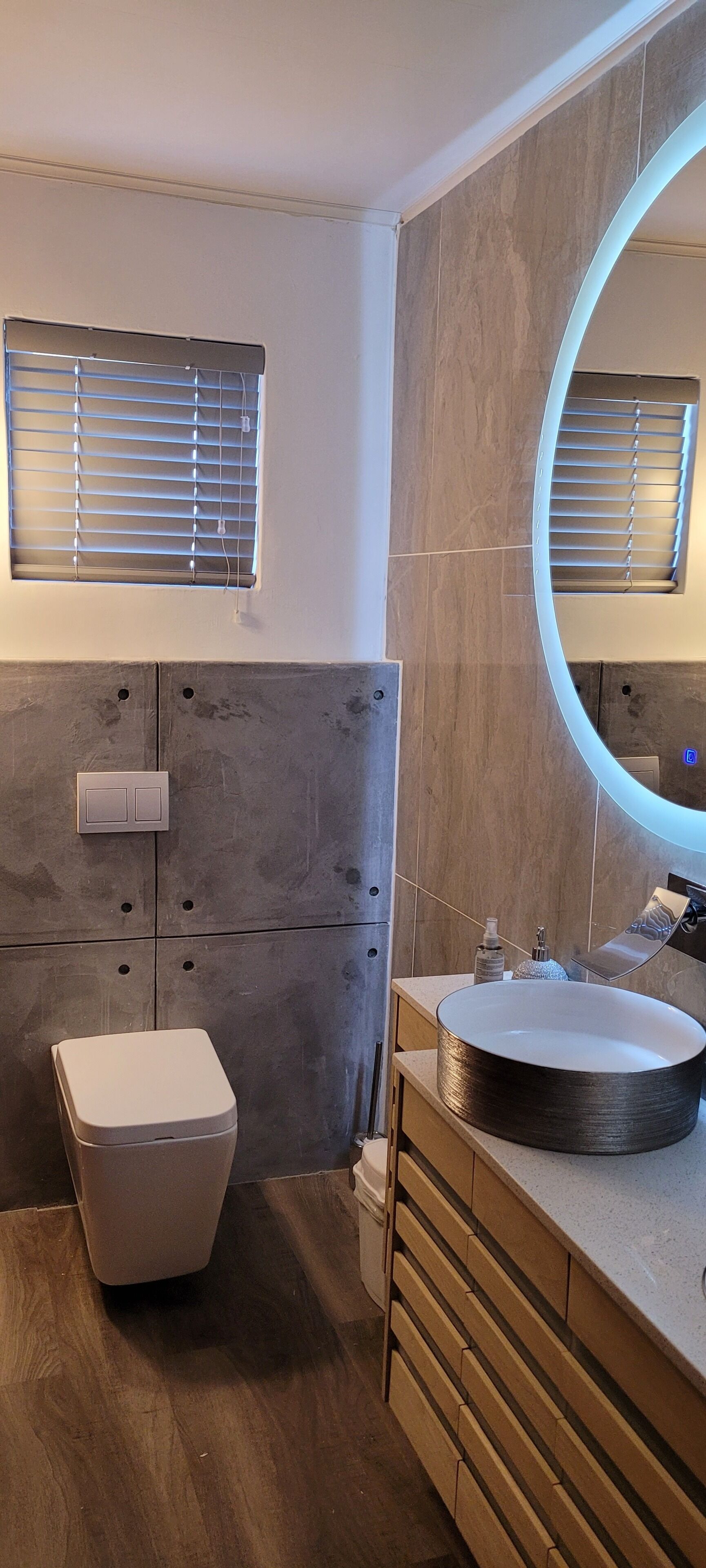 Un baño moderno con un lavabo redondo, un inodoro y un mueble de tocador con cajones. Hay una ventana con persianas y un espejo con luz.