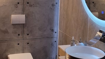 Un baño moderno con un lavabo redondo, un inodoro y un mueble de tocador con cajones. Hay una ventana con persianas y un espejo con luz.