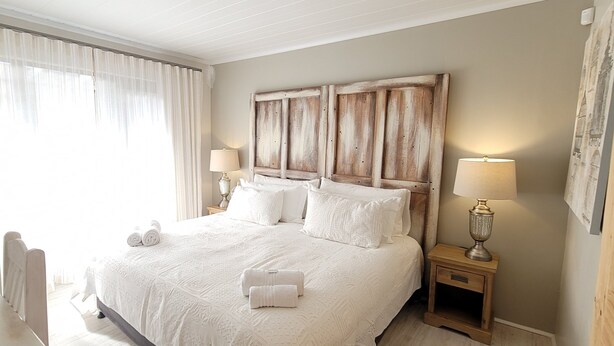 A neatly made bed with white linens and pillows, a wooden headboard, two bedside lamps, and a window with sheer curtains. - Rox and Sea Country Lodge (Langebaan)