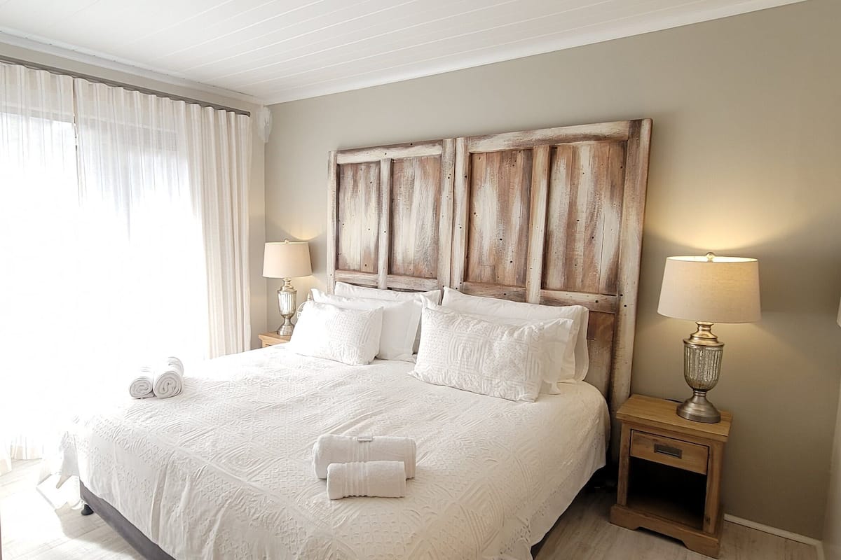 A neatly made bed with white linens and pillows, a wooden headboard, two bedside lamps, and a window with sheer curtains.