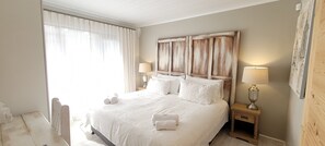 A neatly made bed with white linens and pillows, a wooden headboard, two bedside lamps, and a window with sheer curtains. - Rox and Sea Country Lodge (Langebaan)