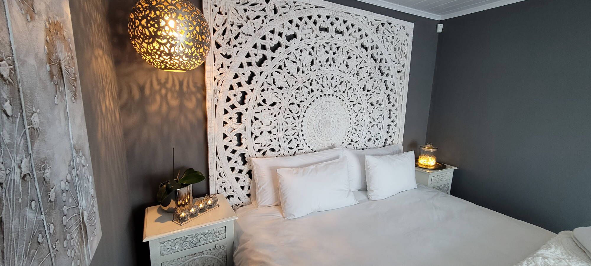 Quarto com cama branca, cabeceira decorativa, mesa de cabeceira com abajur e luminária na parede.
