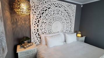 Quarto com cama branca, cabeceira decorativa, mesa de cabeceira com abajur e luminária na parede.