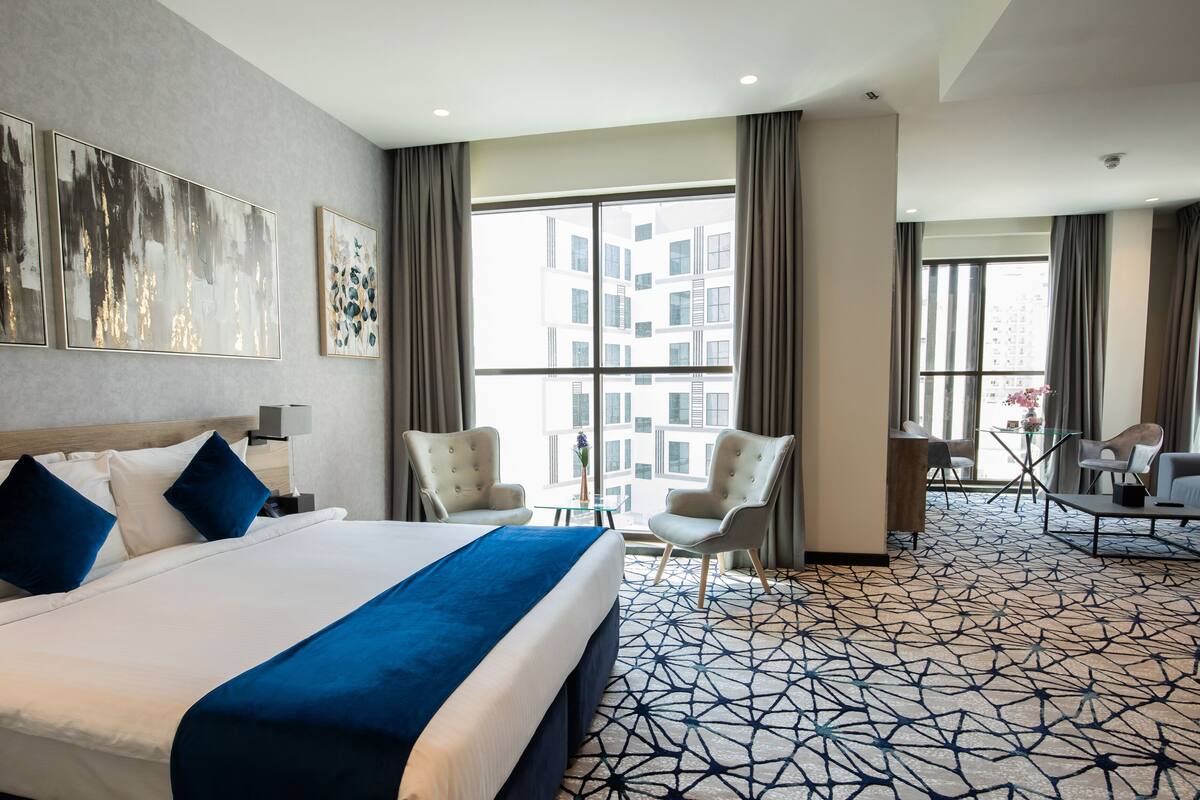 Une chambre d’hôtel avec un grand lit, deux fauteuils, une petite table et une vue sur la ville grâce à de grandes fenêtres.
