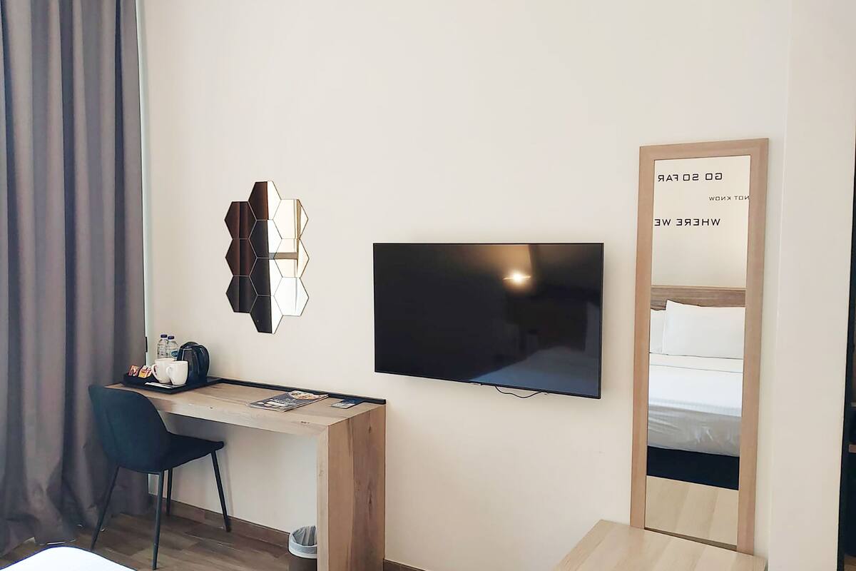Une chambre d’hôtel avec un bureau en bois, un téléviseur à écran plat, un miroir et un lit doté d’une tête de lit blanche.