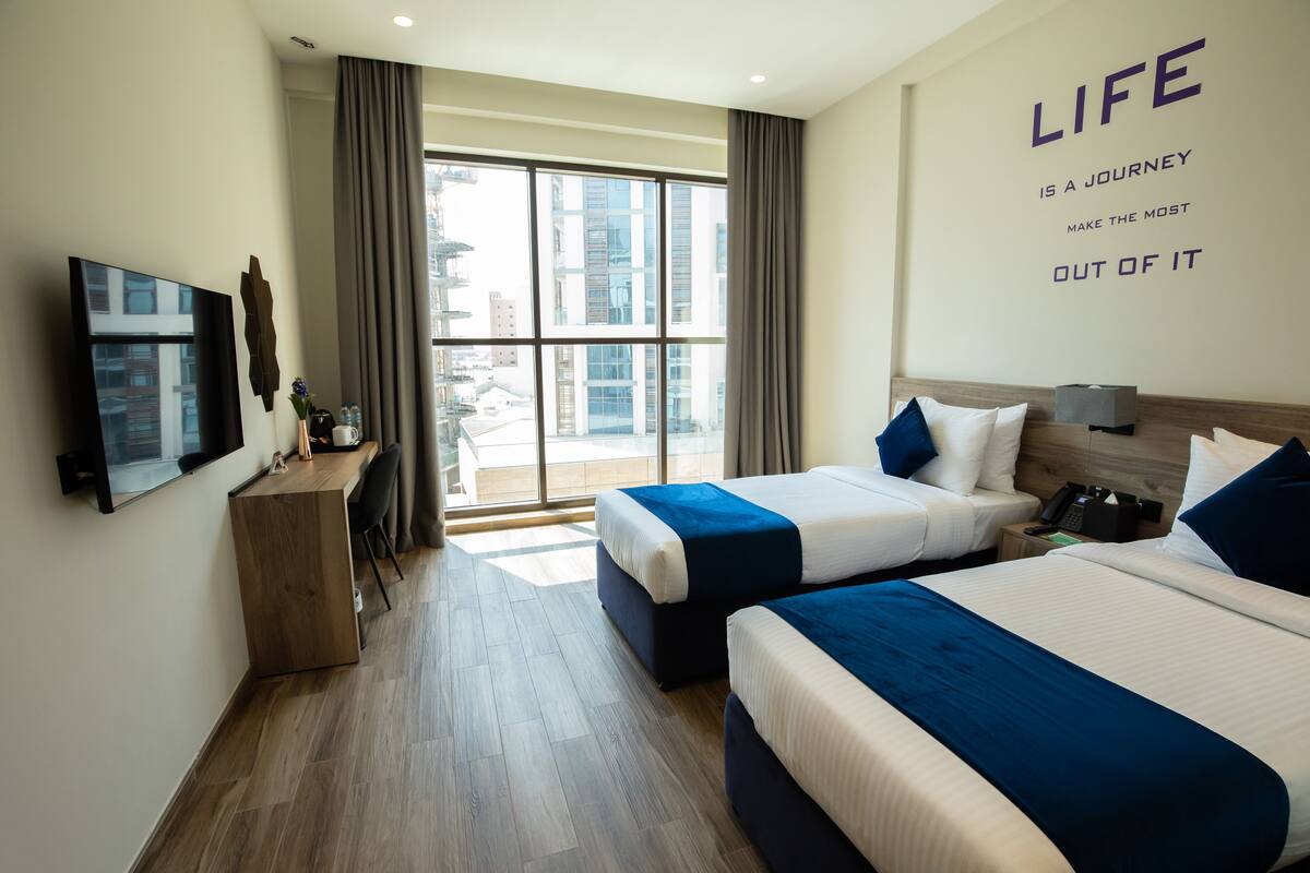 Une chambre d’hôtel avec deux lits, un bureau, une télévision et une grande fenêtre avec des rideaux.