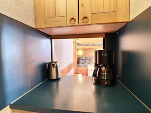 A kitchenette with a coffee maker and a kettle on a blue countertop, wooden cabinets above, and a view into a room with patterned tiles.