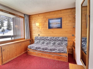 A cozy room with a wooden wall, a window with a view of a snowy landscape, a bed with patterned bedding, and a framed picture of a coastal scene.