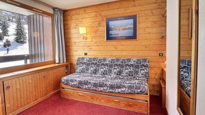 Una habitación acogedora con pared de madera, ventana con vistas a un paisaje nevado, cama con ropa de cama estampada y un cuadro con una escena costera.