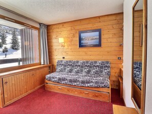 Ein gemütliches Zimmer mit einer Holzwand, einem Fenster mit Blick auf eine verschneite Landschaft, einem Bett mit gemusterten Betttextilien und einem gerahmten Bild einer Küstenansicht.