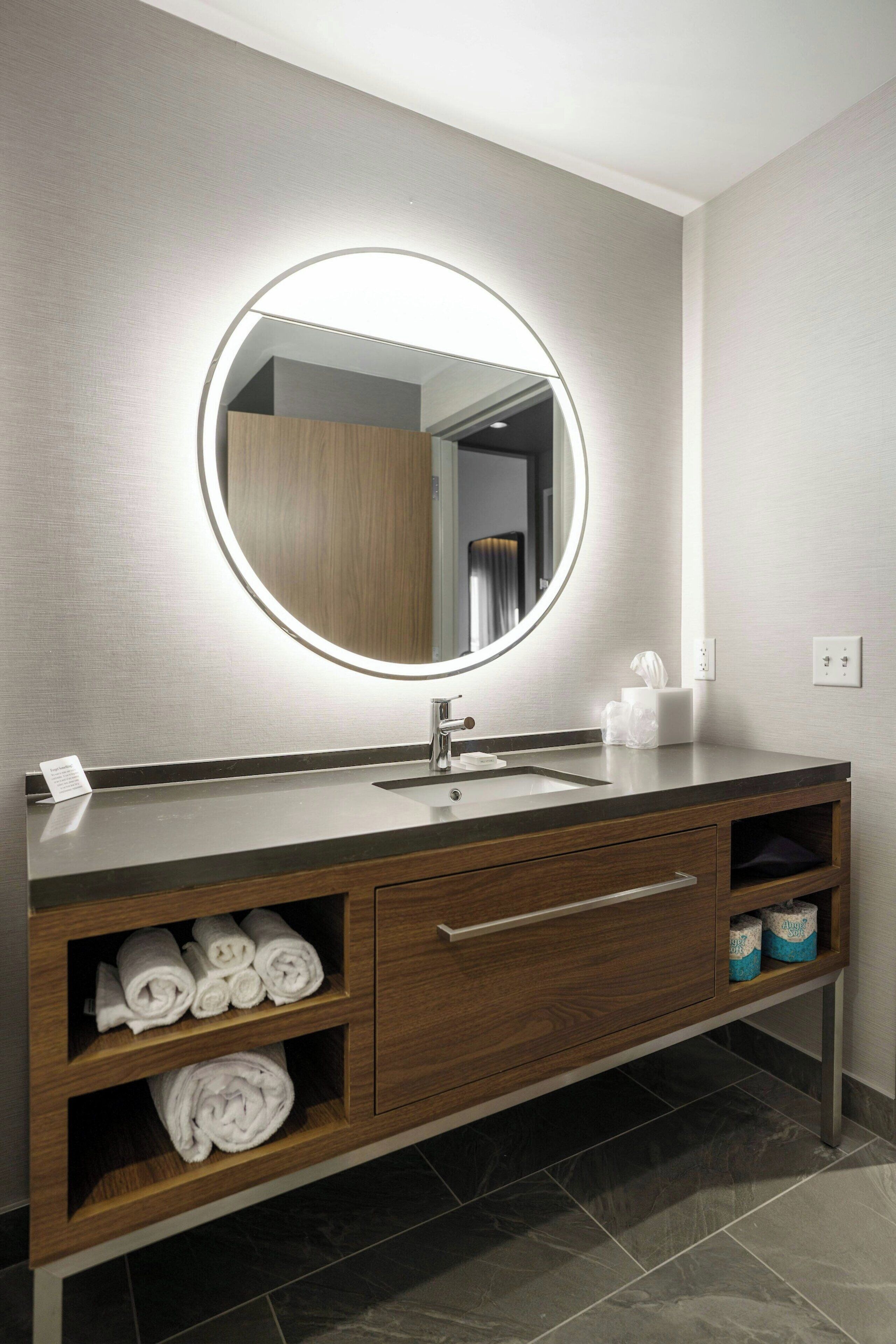 A modern bathroom with a round mirror, a sink, and wooden vanity with shelves for towels.