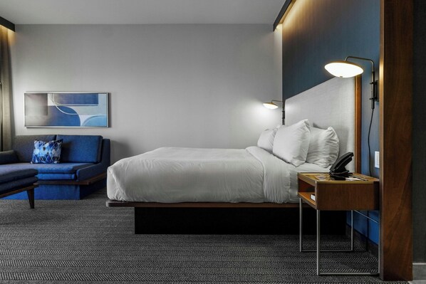 A hotel room with a bed, a bedside table with a lamp, a chair, a sofa, and a wall-mounted abstract painting. - Courtyard by Marriott Indianapolis Fishers (Fishers)