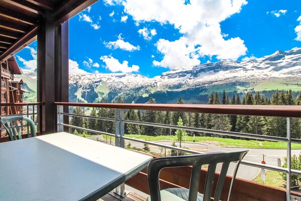 Un balcon avec une table et des chaises, donnant sur un paysage montagneux aux sommets enneigés et sous un ciel bleu dégagé.