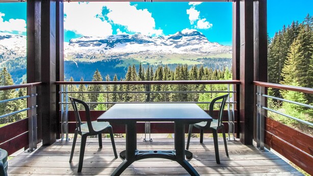 A balcony with a table and chairs, overlooking a mountainous landscape with snow-capped peaks and dense forests. - South-facing newly renovated 2-bed apartment (Flaine)