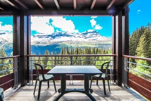 Een balkon met een tafel en stoelen, met uitzicht op een bergachtig landschap met besneeuwde toppen en dichte bossen.