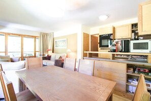A dining area with a wooden table and chairs, a kitchen with wooden cabinets and appliances, and a view through large windows.