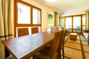 A dining area with a wooden table and chairs, large windows with curtains, and a view of the outdoors.