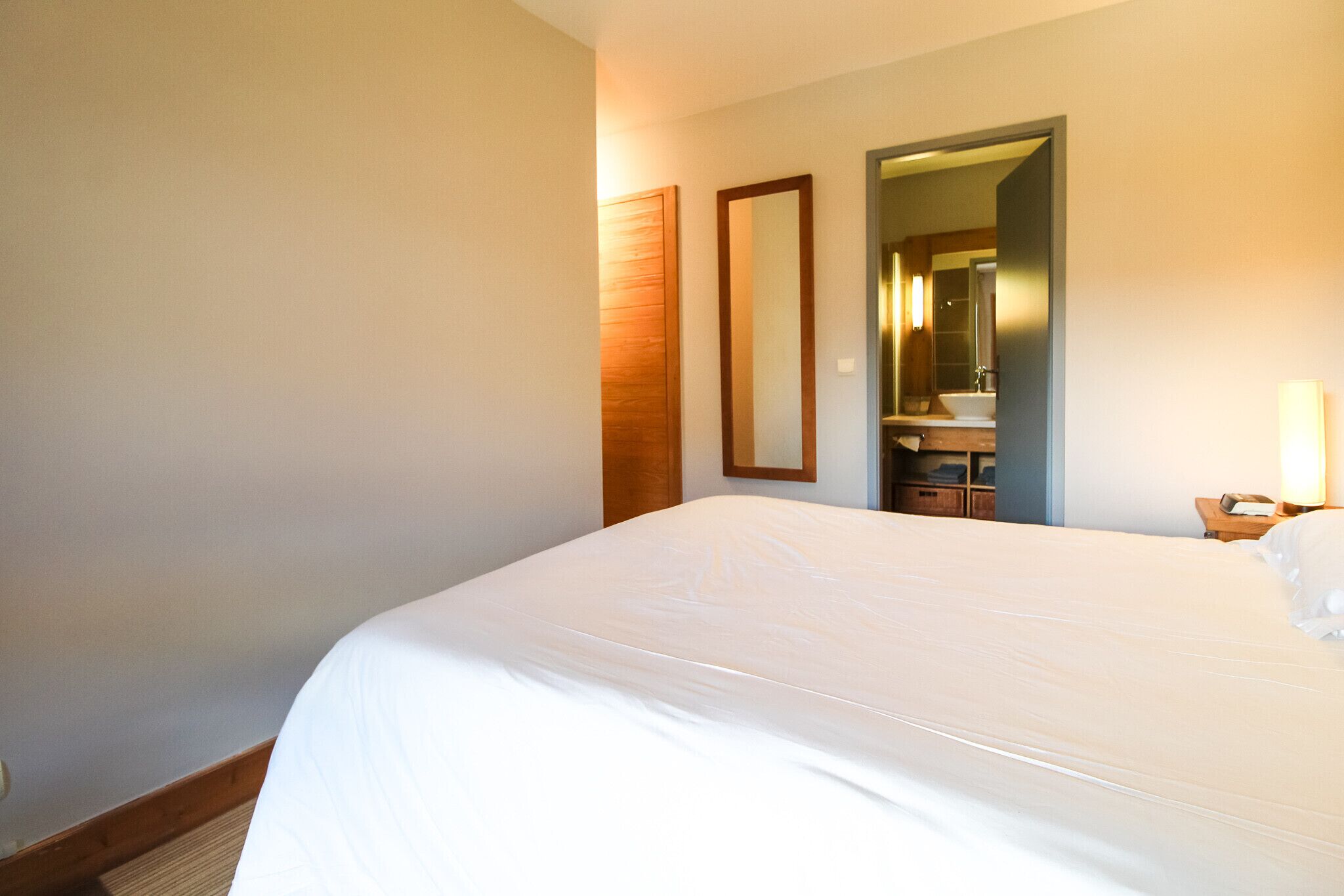 Habitación de hotel con una cama grande, cabecera de madera, un espejo, una mesita de noche y un baño visible a través de una puerta abierta.