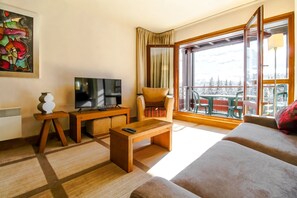A modern living room with a sofa, a TV, a wooden coffee table, and a balcony with a view of mountains.