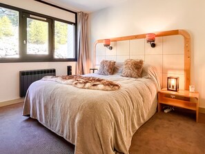 Une chambre avec un grand lit, une table de chevet avec une lampe, une fenêtre avec des rideaux et un radiateur.