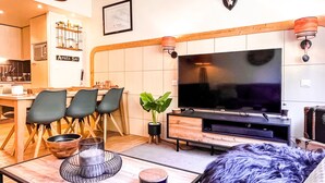 Ein modernes Wohnzimmer mit Flachbildfernseher, einem Essbereich mit Holzkästen und einer Küche im Hintergrund.