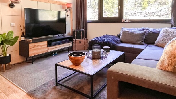 Ein modernes Wohnzimmer mit Flachbildfernseher, Holzwandverkleidung, einem großen Fenster mit Vorhängen, einer Couch und einem Couchtisch.