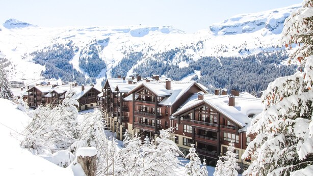 Une station de montagne enneigée, avec des bâtiments de style chalet, une piste de ski et des collines boisées.
