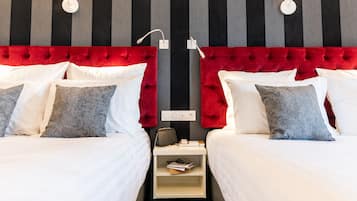 Habitación de hotel con dos camas, cada una con ropa blanca y almohadas grises estampadas. Tiene un cabecero tapizado rojo, una mesita de noche blanca con lámpara y una lámpara de pared.